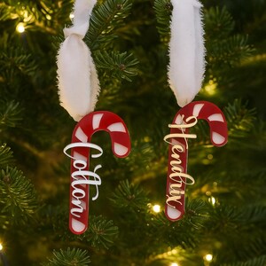 May include: Two candy cane Christmas ornaments hanging on a tree. The ornaments are red and white with the names "Cotton" and "Hendrix" in cursive script. Each ornament is suspended by a white ribbon.