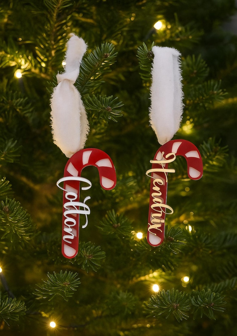 May include: Two candy cane Christmas ornaments hanging on a tree. The ornaments are red and white with the names "Cotton" and "Hendrix" in cursive script. Each ornament is suspended by a white ribbon.