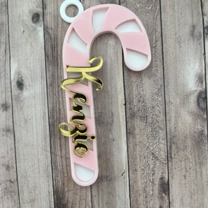 May include: A pink and white candy cane ornament with the name "Kenzie" in gold script. The ornament has a white loop at the top for hanging. The candy cane design features alternating pink and white stripes.