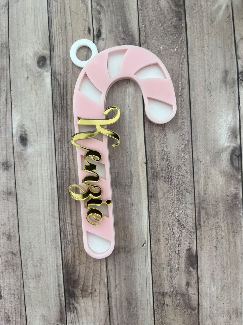May include: A pink and white candy cane ornament with the name "Kenzie" in gold script. The ornament has a white loop at the top for hanging. The candy cane design features alternating pink and white stripes.