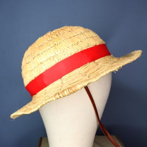 May include: A straw hat with a wide brim and a vibrant red band. The hat is displayed on a white mannequin head, with a brown chin strap. The hat's woven texture and bright colors make it a stylish accessory.