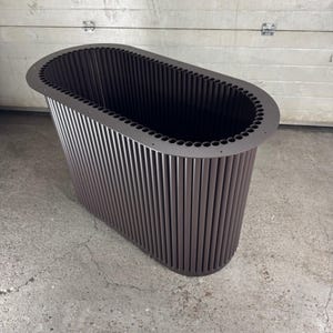 Könnte beinhalten: Ein brauner, oval geformter Pflanzkübel mit vertikal gerippten Seiten. Der obere Rand hat eine Reihe von gleichmäßig verteilten Löchern. Der Pflanzkübel ist aus Metall und steht auf einer Betonoberfläche. Das Design ist modern und minimalistisch.