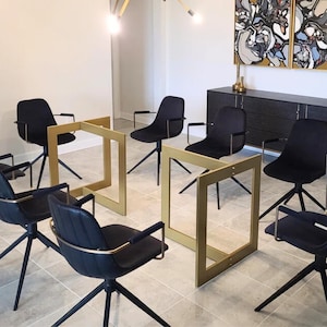 Könnte beinhalten: Innenaufnahme eines modernen Esszimmersets. Das Set umfasst schwarze Drehstühle mit Armlehnen und goldfarbene, geometrische Tischrahmen. Die Stühle sind auf einem gefliesten Boden um die Rahmen angeordnet, mit einem dunklen Schrank und Kunstwerken im Hintergrund.