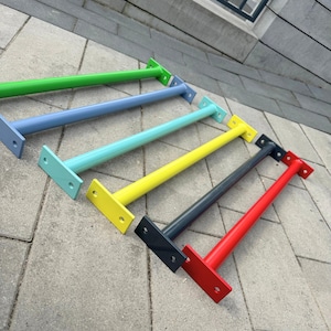 Może przedstawiać: Kolekcja sześciu kolorowych wsporników półkowych. Wsporniki pomalowane są w odcieniach zieleni, błękitu, turkusu, żółci, szarości i czerwieni. Każdy wspornik ma prostokątną płytę montażową i cylindryczne ramię podporowe.