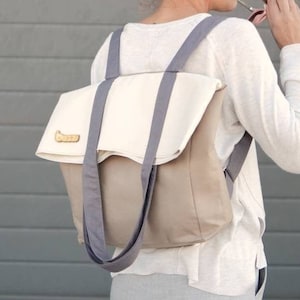 Op de afbeelding: Een vrouw draagt een wit longsleeve shirt en een grijze broek en draagt een grijze en beige canvas rugzak met een witte flap en een houten label.