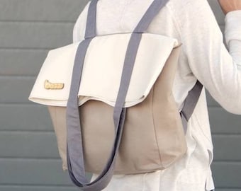 Puede incluir: Una mujer con una camisa blanca de manga larga y pantalones grises lleva una mochila de lona beige y gris con una solapa blanca y una etiqueta de madera.