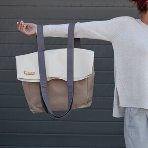 Op de afbeelding: Een grote, beige en bruine tote bag met twee grijze hengsels. De tas heeft een klep die over de bovenkant vouwt en wordt vastgezet met een knoop.