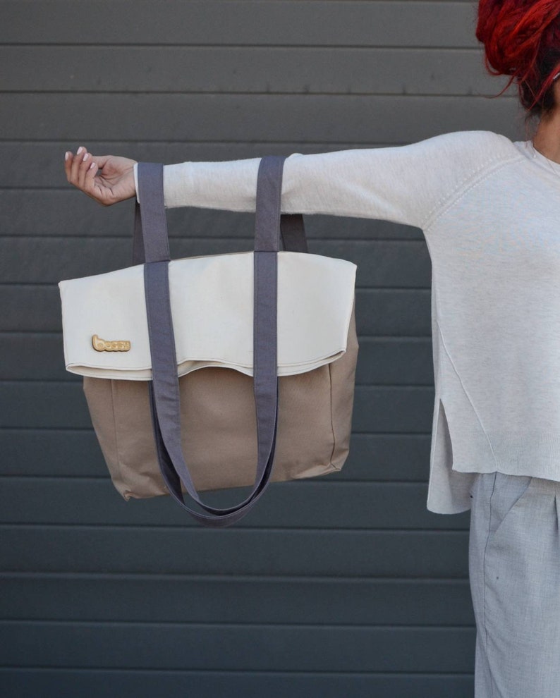 Op de afbeelding: Een grote, beige en bruine tote bag met twee grijze hengsels. De tas heeft een klep die over de bovenkant vouwt en wordt vastgezet met een knoop.