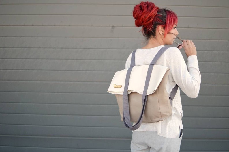 Op de afbeelding: Een vrouw draagt een wit longsleeve shirt en een grijze broek en draagt een grijze en beige canvas rugzak met een witte flap en een houten label.