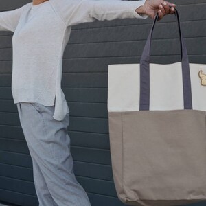 Op de afbeelding: Een grote, tweekleurige tote bag met een witte bovenkant en een bruine onderkant. De tas heeft twee donkergrijze hengsels en een klein houten olifantenbedeltje aan de voorkant.