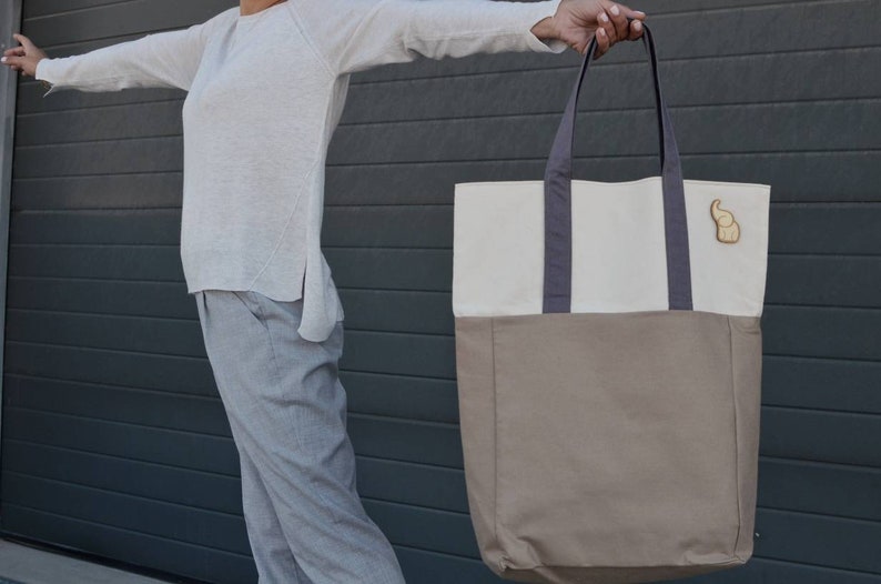 Op de afbeelding: Een grote, tweekleurige tote bag met een witte bovenkant en een bruine onderkant. De tas heeft twee donkergrijze hengsels en een klein houten olifantenbedeltje aan de voorkant.