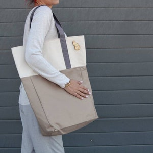 Op de afbeelding: Een grote beige en witte tote bag met een grijze handvat en een kleine olifant charme. De tas is gemaakt van canvas en heeft een ritssluiting.