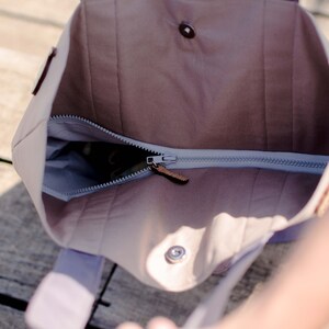 Op de afbeelding: Een lichtbruine canvas tote bag met een ritssluiting en een bruine leren rits. De tas is open en de binnenkant is gevoerd met een lichtblauwe stof.