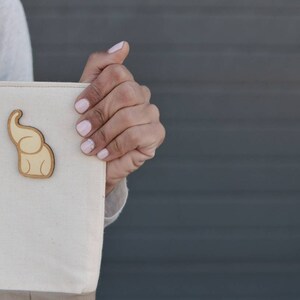 Op de afbeelding: Een beige canvas tote bag met een bruin olifant ontwerp. De tas heeft een grijze band en wordt vastgehouden door een hand.