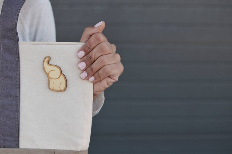 Op de afbeelding: Een beige canvas tote bag met een bruin olifant ontwerp. De tas heeft een grijze band en wordt vastgehouden door een hand.