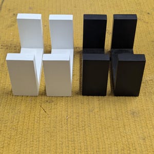 May include: Four wooden wall hooks, two in white and two in black, arranged in a row. Each hook has a rectangular base and a perpendicular arm, creating an L-shape. The hooks are displayed on a yellow surface.