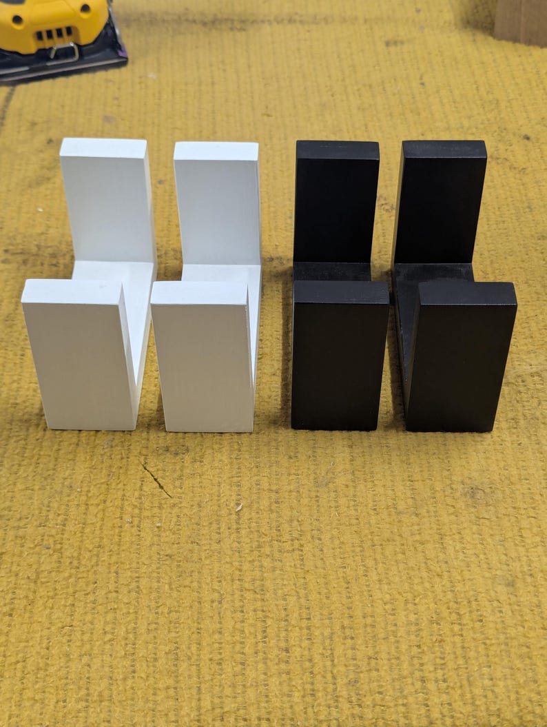 May include: Four wooden wall hooks, two in white and two in black, arranged in a row. Each hook has a rectangular base and a perpendicular arm, creating an L-shape. The hooks are displayed on a yellow surface.