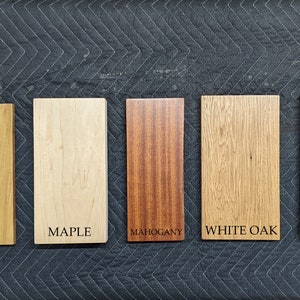 May include: Five rectangular wood samples labeled Poplar, Maple, Mahogany, White Oak, and Black Walnut. The wood samples vary in color from light tan to dark brown. The text labels are in a simple sans-serif font.