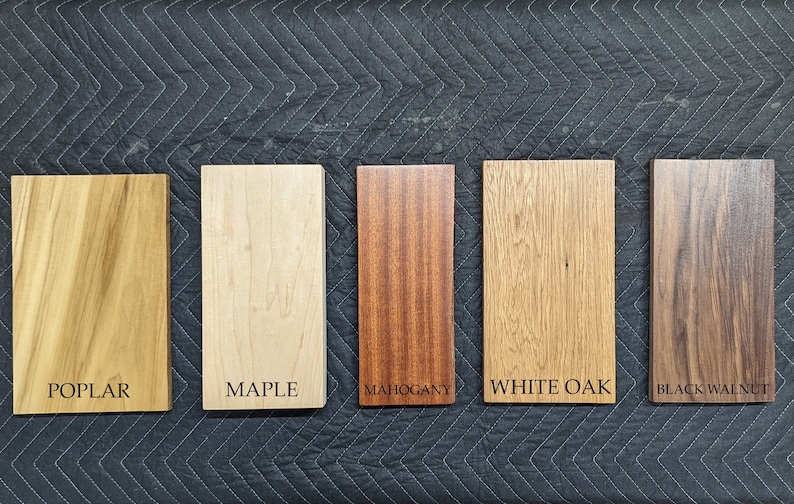 May include: Five rectangular wood samples labeled Poplar, Maple, Mahogany, White Oak, and Black Walnut. The wood samples vary in color from light tan to dark brown. The text labels are in a simple sans-serif font.
