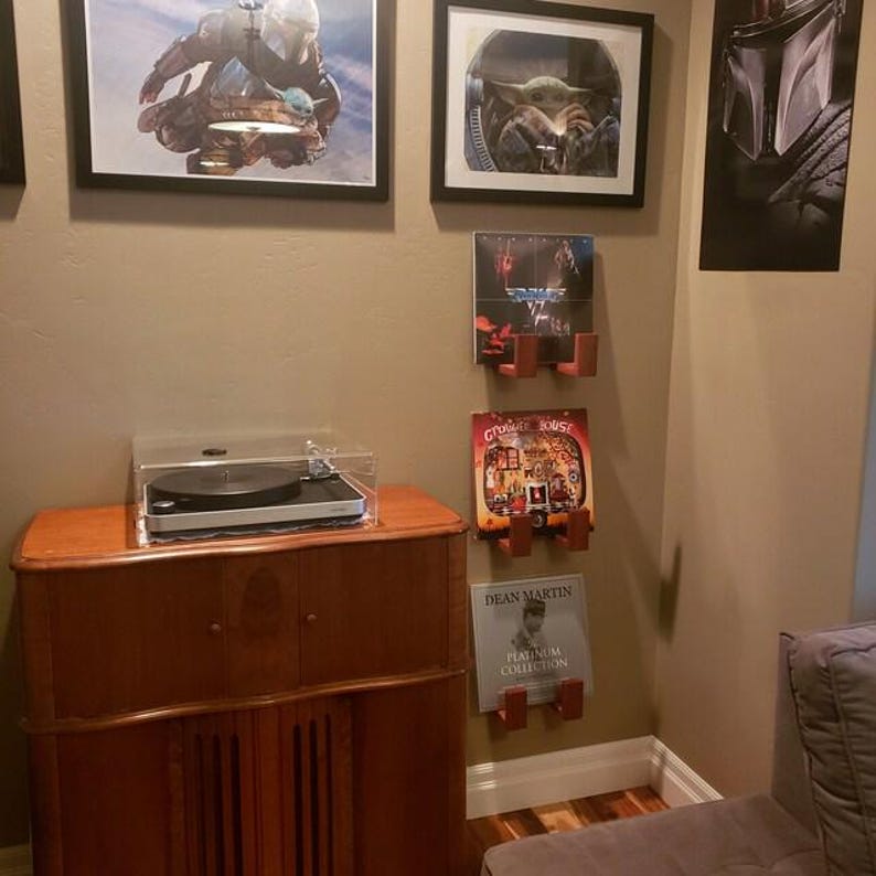 May include: A wooden cabinet with a record player on top, protected by a clear cover. Framed Star Wars art hangs on the wall, along with record holders displaying album covers. The cabinet is a warm brown color.