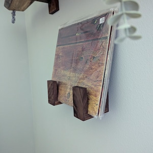 May include: A dark wood record holder mounted on a light gray wall, displaying a vinyl record album. The album cover is visible, featuring a map-like design with text. A wooden shelf is above the record holder. Decorative plant leaves are in the upper right corner.
