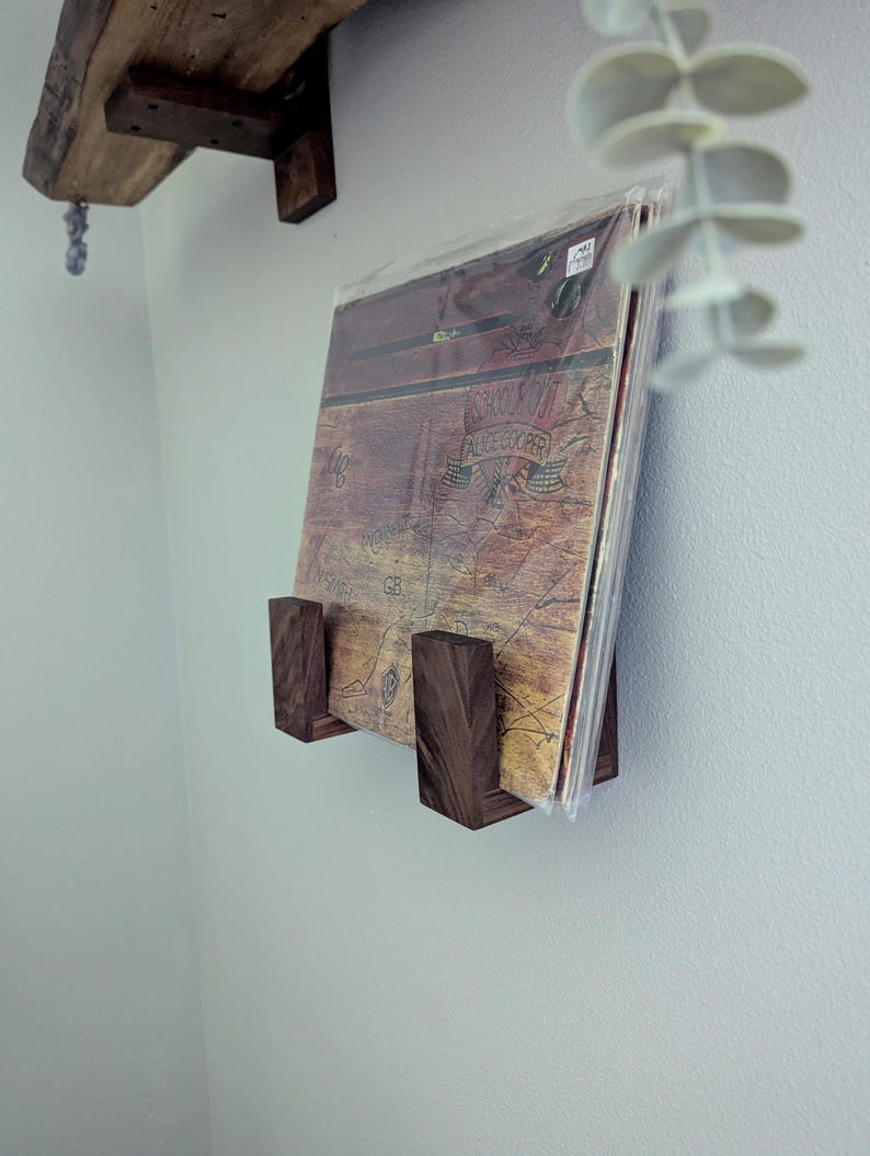 May include: A dark wood record holder mounted on a light gray wall, displaying a vinyl record album. The album cover is visible, featuring a map-like design with text. A wooden shelf is above the record holder. Decorative plant leaves are in the upper right corner.
