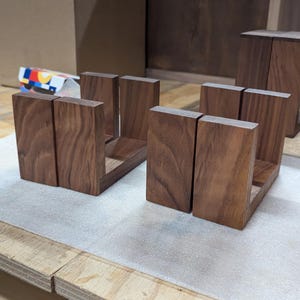 May include: Two sets of wooden bookends, each composed of two vertical pieces joined by a horizontal base. The bookends are made of dark brown wood and are displayed on a light-colored surface.