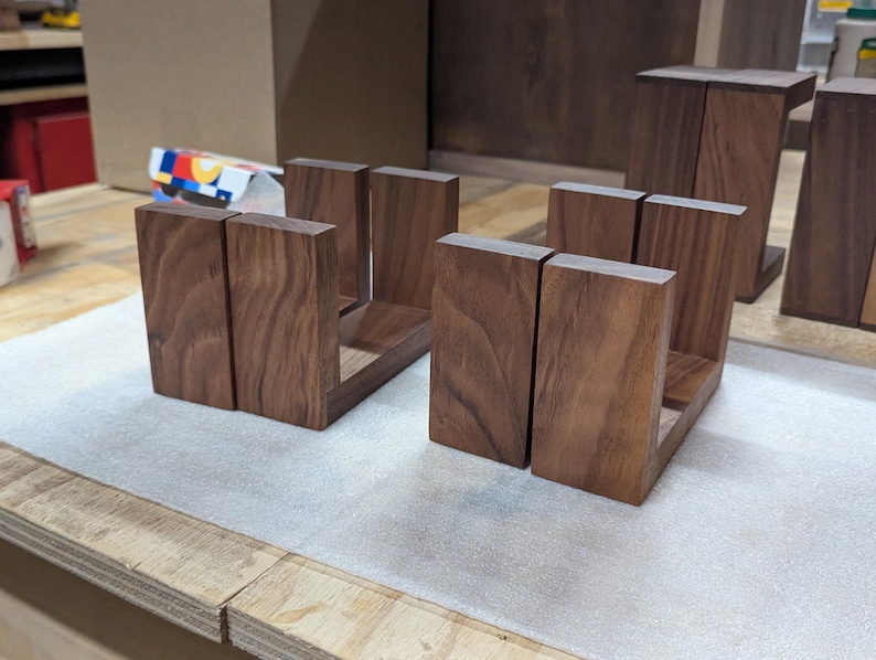 May include: Two sets of wooden bookends, each composed of two vertical pieces joined by a horizontal base. The bookends are made of dark brown wood and are displayed on a light-colored surface.