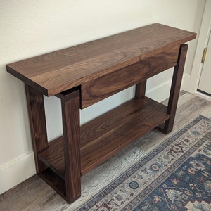 May include: A dark brown wooden console table with a single drawer and a lower shelf. The table has a simple, modern design and is made of solid wood.
