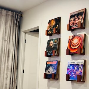 May include: Six vinyl record albums displayed on a white wall in wooden holders. The album covers feature various designs, including portraits, abstract art, and text. The holders are a dark brown color.