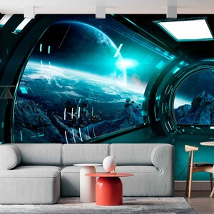 Op de afbeelding: Een futuristisch ruimtethema-muurschildering met een uitzicht op een planeet vanuit een ruimteschipraam. De sc&egrave;ne wordt gedomineerd door tinten blauw en turquoise, met een gedetailleerde weergave van een planeet en een bergachtig landschap. De kamer omvat een grijze bank, een rode bijzettafel en een houten eethoek.