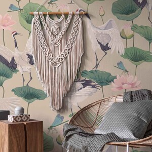 May include: A beige wall with a floral wallpaper featuring pink lotus flowers, green leaves, and white cranes. A white macrame wall hanging is centered on the wall. A wicker chair with a gray cushion sits in front of the wall.