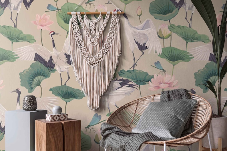 May include: A beige wall with a floral wallpaper featuring pink lotus flowers, green leaves, and white cranes. A white macrame wall hanging is centered on the wall. A wicker chair with a gray cushion sits in front of the wall.