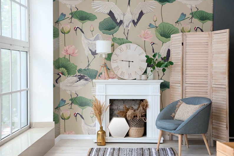 May include: A white fireplace mantel with a white clock on top. The clock has roman numerals and black hands. The fireplace has a brick interior and a white surround. There is a blue armchair with a beige pillow in front of the fireplace. The wall behind the fireplace has a wallpaper with a green and pink floral pattern and white cranes.