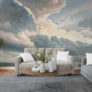 May include: A living room interior with a light grey sofa and a coffee table with a vase of green plants and decorative objects. The wall behind the sofa is decorated with a large painting of a cloudy sky in shades of blue, grey, and white.