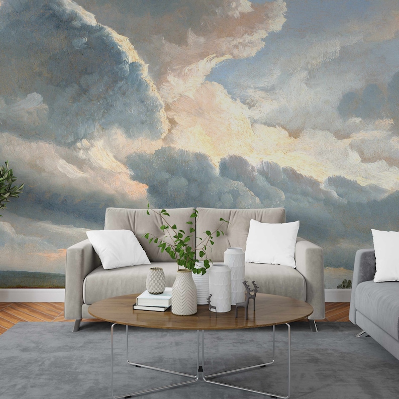 May include: A living room interior with a light grey sofa and a coffee table with a vase of green plants and decorative objects. The wall behind the sofa is decorated with a large painting of a cloudy sky in shades of blue, grey, and white.