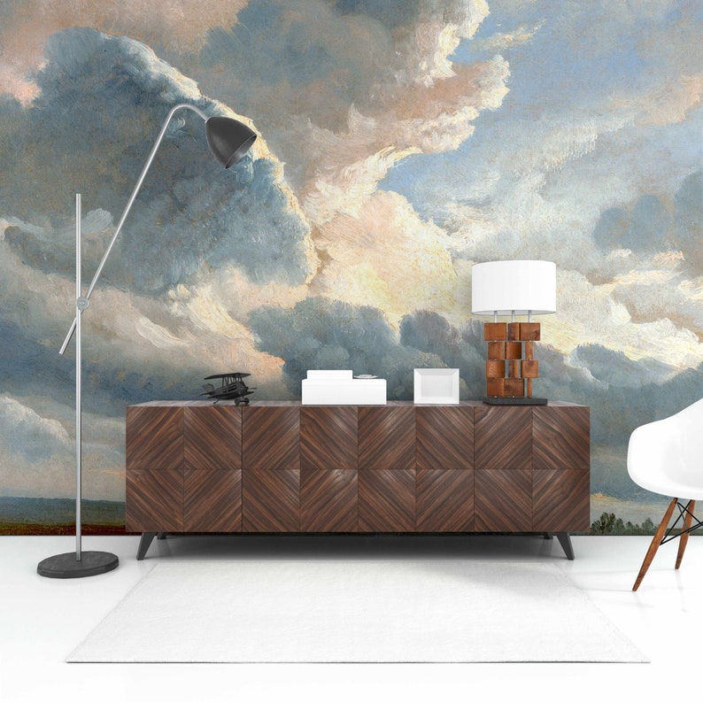 May include: A dark wood cabinet with a geometric pattern on the front sits in front of a wall mural depicting a cloudy sky. A lamp with a white shade and a silver floor lamp are on top of the cabinet. A white chair is in the foreground.