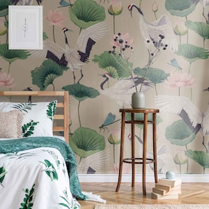 May include: A bedroom with a beige wall featuring a wallpaper with a crane and lotus flower design. The bed has a white duvet with green leaves and a green throw blanket. A wooden stool with a vase sits in front of the bed.