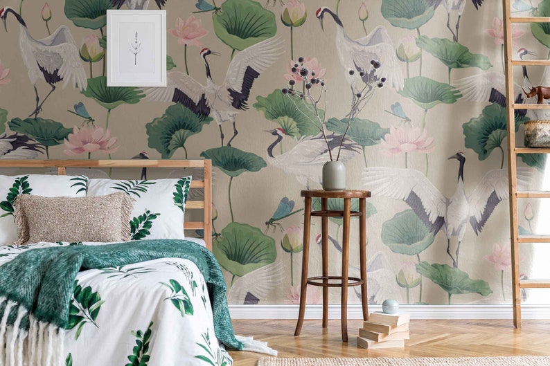 May include: A bedroom with a beige wall featuring a wallpaper with a crane and lotus flower design. The bed has a white duvet with green leaves and a green throw blanket. A wooden stool with a vase sits in front of the bed.