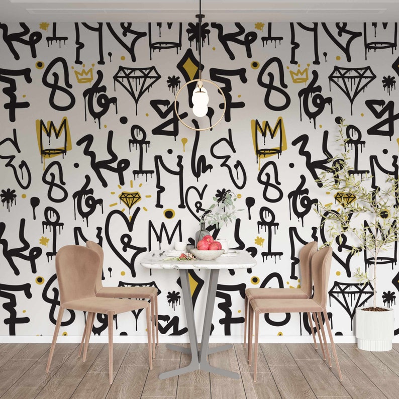 May include: A dining room with a white wall featuring a black and gold graffiti pattern. The room has a square table with three chairs and a potted plant.