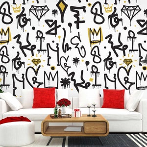 May include: A white couch with red pillows sits in front of a wall with a black and gold graffiti pattern. The pattern includes crowns, diamonds, hearts, and other abstract shapes. A coffee table with a wooden top and black legs sits in front of the couch. A small plant is in a white pot on the left side of the image.