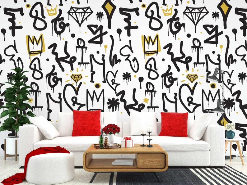 May include: A white couch with red pillows sits in front of a wall with a black and gold graffiti pattern. The pattern includes crowns, diamonds, hearts, and other abstract shapes. A coffee table with a wooden top and black legs sits in front of the couch. A small plant is in a white pot on the left side of the image.