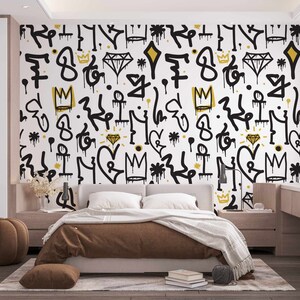 May include: A bedroom with a white wall featuring a black and gold graffiti pattern. The wall is decorated with crowns, diamonds, and other abstract shapes. A brown bed with a brown blanket and pillows is in front of the wall. A brown ottoman is in the foreground.