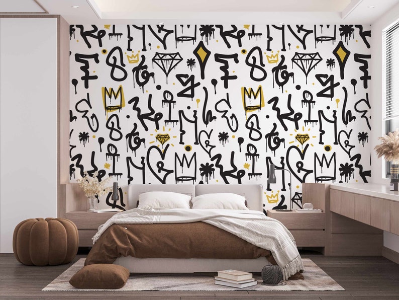 May include: A bedroom with a white wall featuring a black and gold graffiti pattern. The wall is decorated with crowns, diamonds, and other abstract shapes. A brown bed with a brown blanket and pillows is in front of the wall. A brown ottoman is in the foreground.