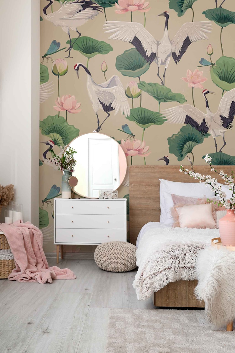 May include: A bedroom with a white dresser, a bed with a white fluffy blanket and a pink throw pillow, a round mirror, a woven ottoman, and a wall mural featuring cranes, lotus flowers, and dragonflies.