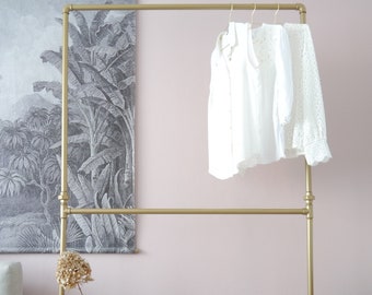 May include: A gold metal clothing rack with two bars. The rack is standing on a wooden floor in front of a pink wall. There are white clothes hanging on the rack.