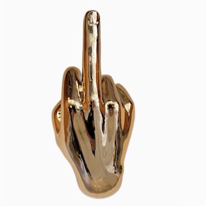 Könnte beinhalten: Eine goldfarbene Handskulptur mit nach oben ausgestrecktem Mittelfinger. Die Skulptur ist hochreflektierend, mit sichtbaren Lichtreflexen auf ihrer Oberfläche. Die Hand ist zu einer Faust geballt, der Daumen ist eingeklappt.