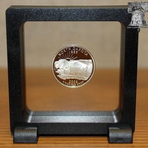 May include: A silver North Dakota quarter coin from 2006, featuring a bison and the words "North Dakota" and "E Pluribus Unum." The coin is displayed in a black, square, floating frame.