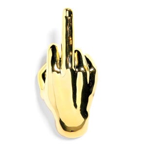 May include: A gold-colored hand sculpture with the middle finger extended upwards. The sculpture is highly reflective and appears to be made of metal. The hand is positioned against a white background.