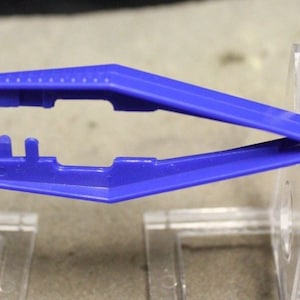 May include: A blue plastic tweezer tool with serrated tips. The tweezer is open, revealing its design. The tool is designed for precision work, likely for crafts or small object handling. The tweezer is made of a durable plastic material.